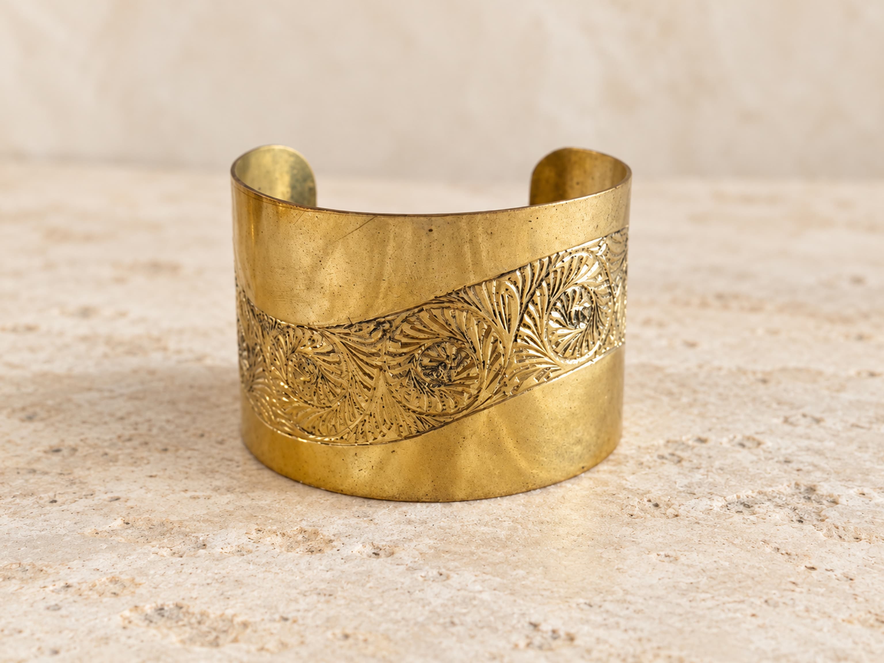 Vista frontal oblicua del brazalete ancho de latón con franja grabada de hojas y espirales y abertura posterior.