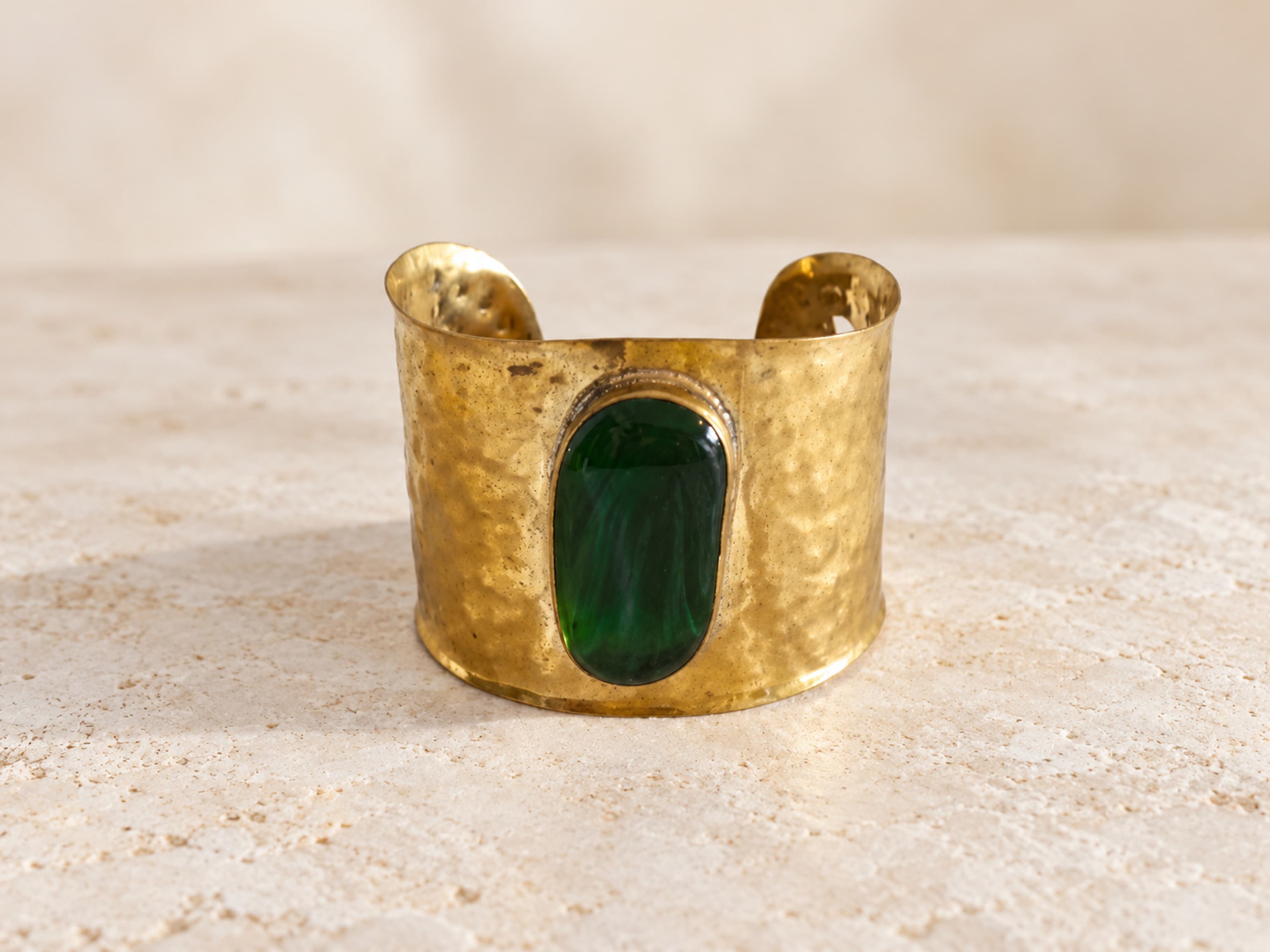 Brazalete ancho y abierto de latón de tono dorado con textura irregular y piedra verde alargada colocada en vertical.