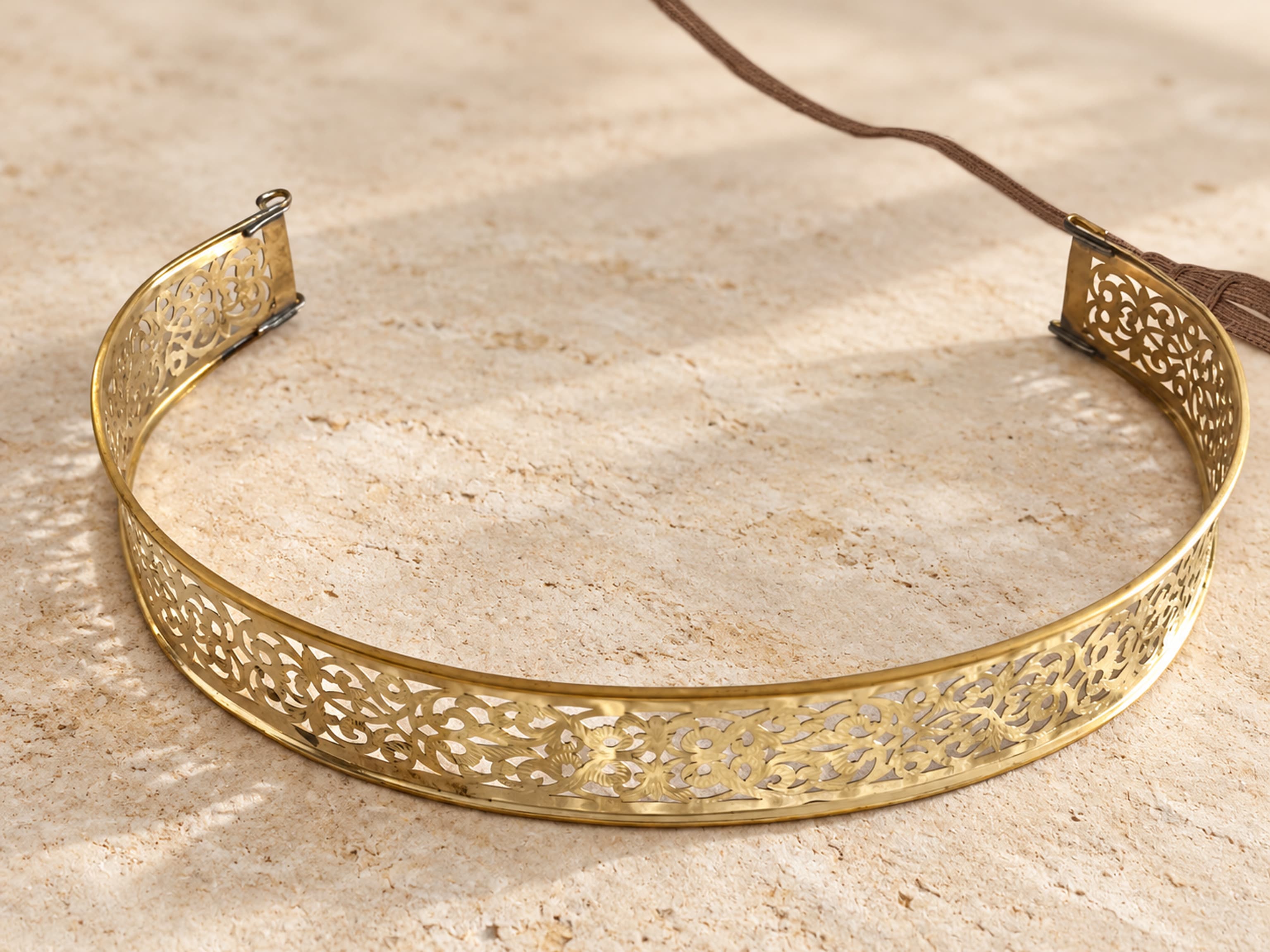 Cinturón estrecho y curvado de latón dorado con calado floral, herrajes en los extremos y cinta marrón.