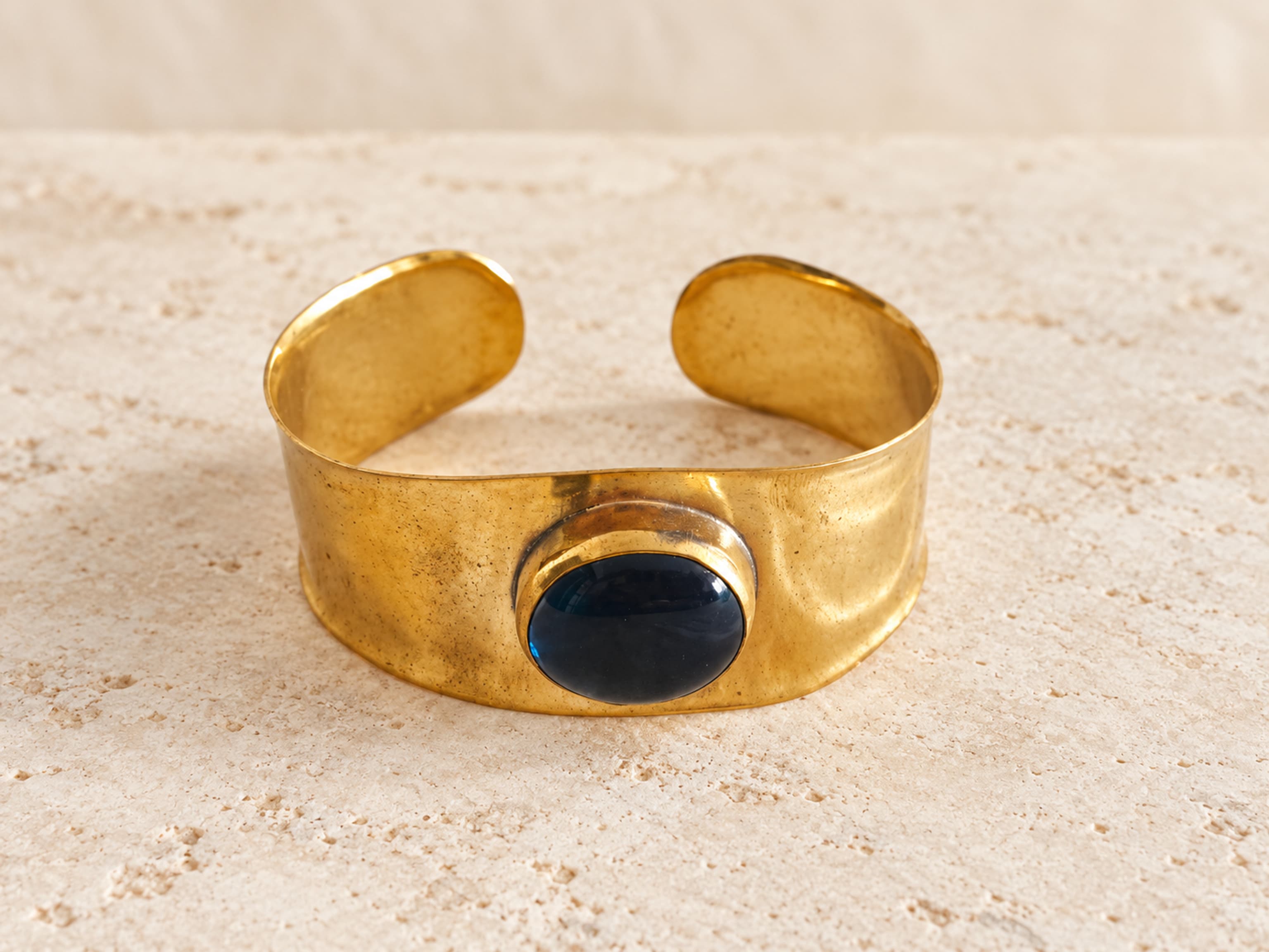 Brazalete ancho y abierto de latón dorado con piedra azul oscuro abombada, vista oblicua.
