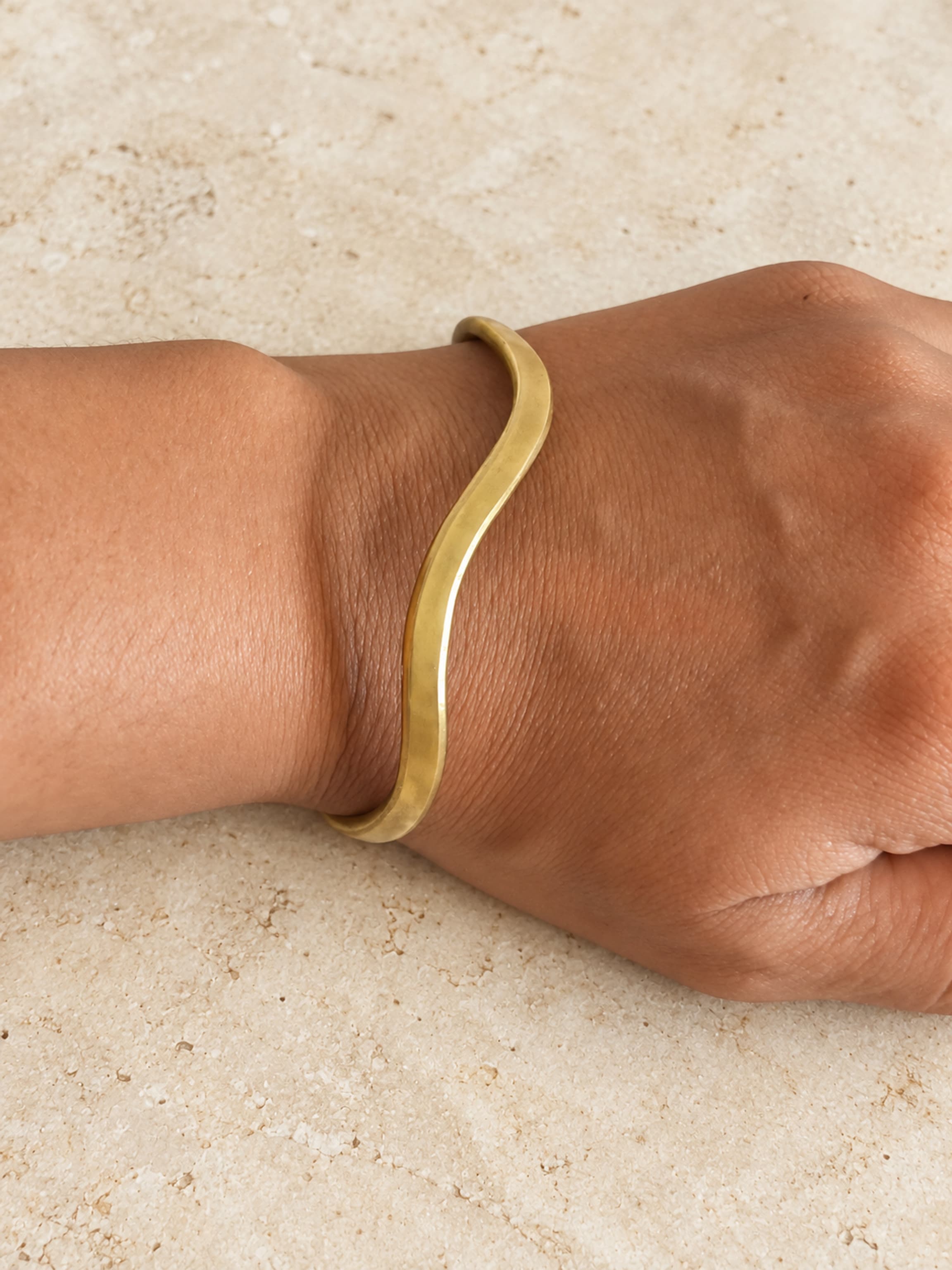 Brazalete fino de latón de tono dorado con forma de ola, llevado en la muñeca.