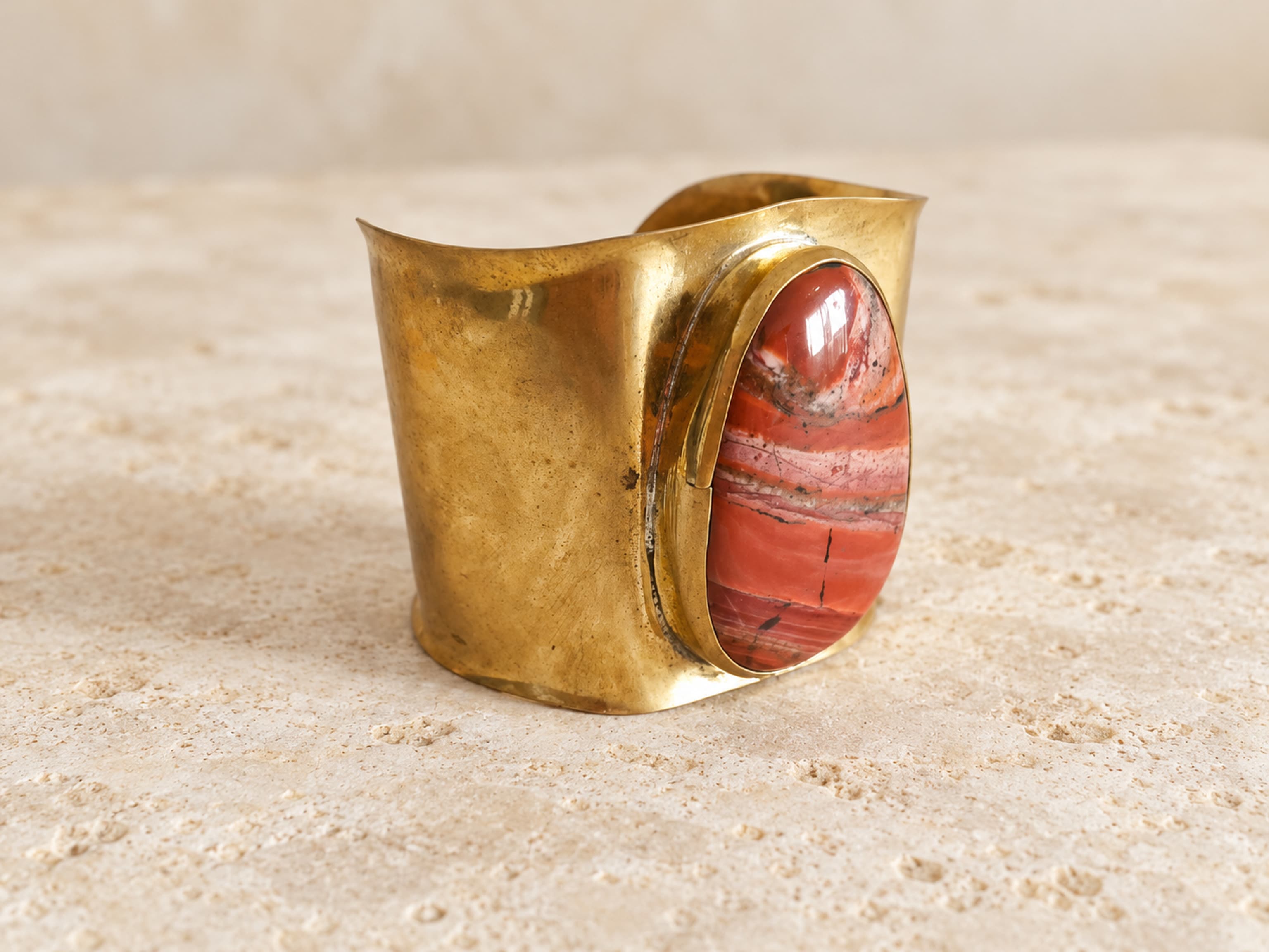 Vista de tres cuartos del brazalete ancho de latón dorado con piedra naranja rojiza alargada y vetas diagonales.